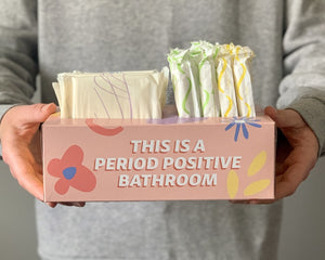 Close up photo of "Your Period Box" on the front it says "This is a period positive bathroom". A person is holding the box up to the camera