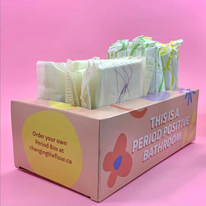 Phot of Your Period Box, with pads and tampons inserted. On teh front of the box it says "This is a period positive bathroom". On the left side it says "Order Your Own Period Box at changingtheflow.ca" in a yellow circle