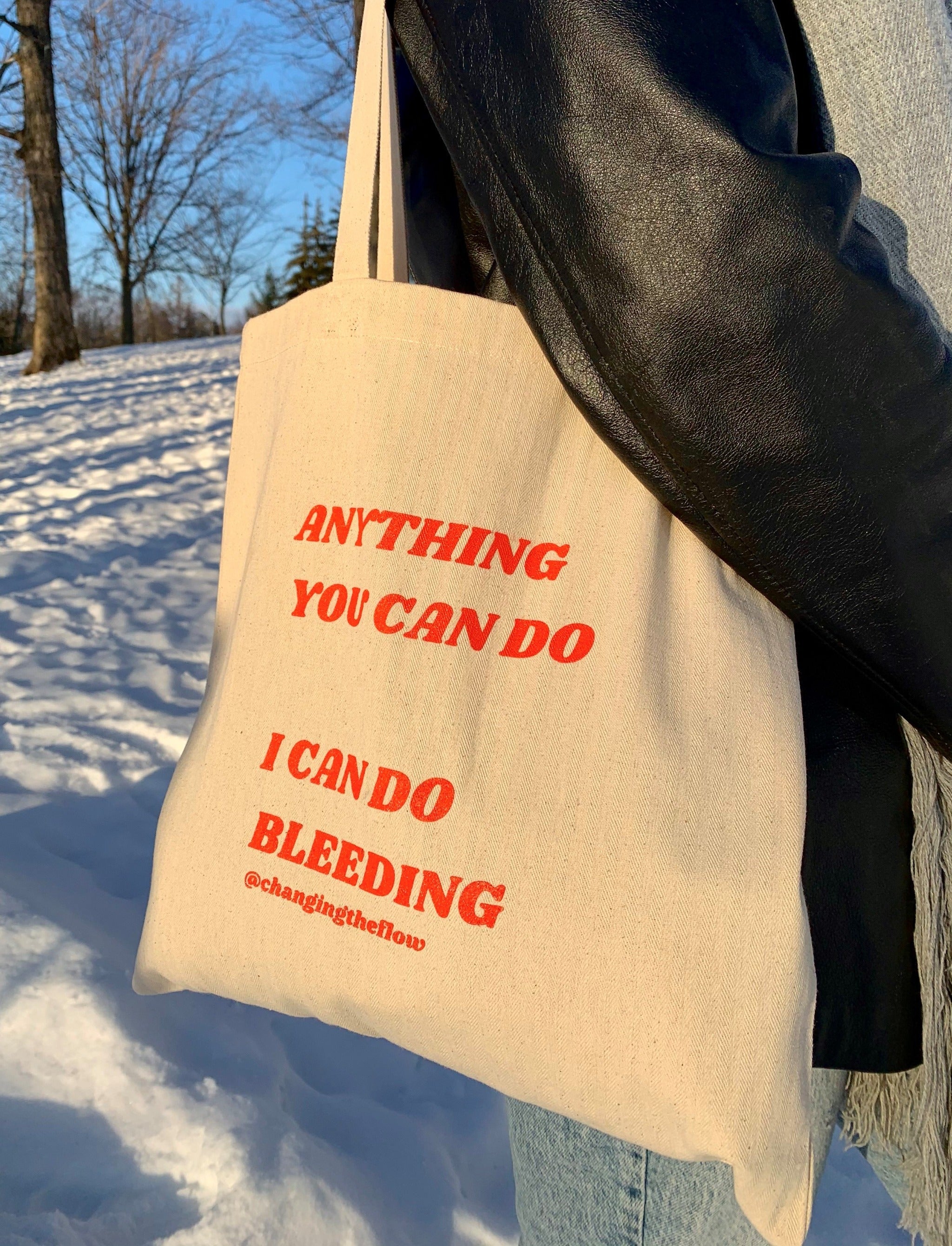 image of our canvas totes that says Anything You Can Do I Can Do Bleeding shown hanging on someone's shoulder, with snow on the ground in the background