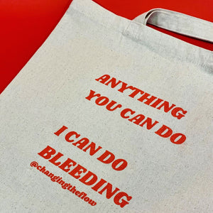 an image of our canvas tote, flatttened on a red background. The writing on the tote is in red and says Anything You Can Do I Can Do Bleeding @changingtheflow