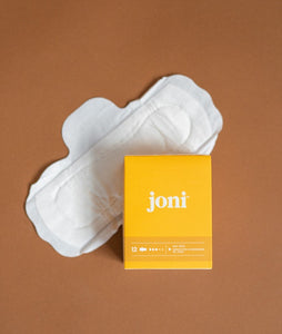 A joni bamboo pad laid out on a light brown background with a yellow box of joni day pads resting on a portion of the pad